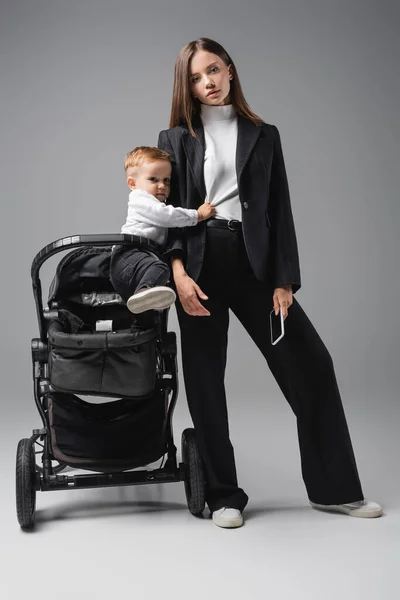 Empresaria sosteniendo smartphone con pantalla en blanco cerca de cochecito con hijo en gris - foto de stock