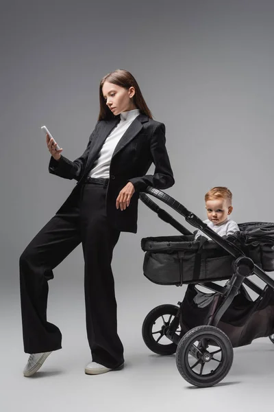 Mujer en traje negro usando teléfono inteligente cerca de cochecito con hijo pequeño en gris - foto de stock