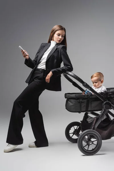 Mujer con smartphone mirando hijo jugando con juguete coche en cochecito en gris - foto de stock
