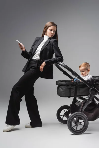 Woman with smartphone looking at son playing with toy car in pram on grey — Stock Photo