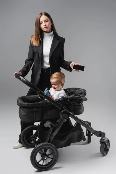 Mujer sosteniendo teléfono inteligente con pantalla en blanco cerca de hijo jugando en carro de bebé en gris - foto de stock