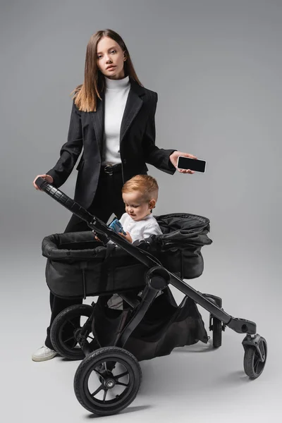 Woman holding smartphone with blank screen near son playing in baby carriage on grey — Stock Photo