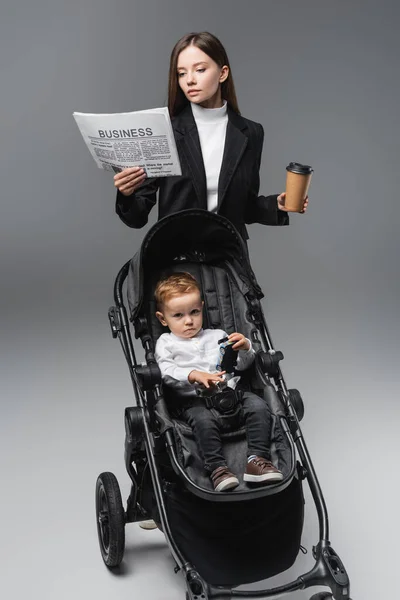 Бізнес-леді з кавою, щоб піти читати газету біля сина в дитячій колясці на сірому — стокове фото