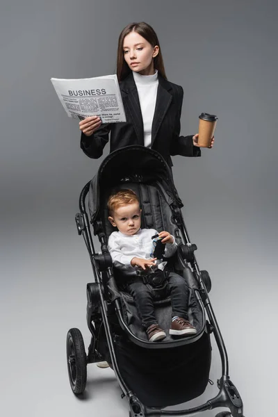 Mujer de negocios con café para ir a leer el periódico cerca de hijo en carro de bebé en gris - foto de stock