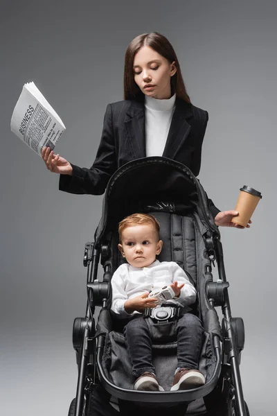 Businesswoman with newspaper and coffee to go near son sitting in pram with toy car isolated on grey — Foto stock