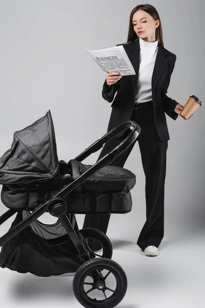Businesswoman reading newspaper while standing with paper cup near pram on grey — Stock Photo
