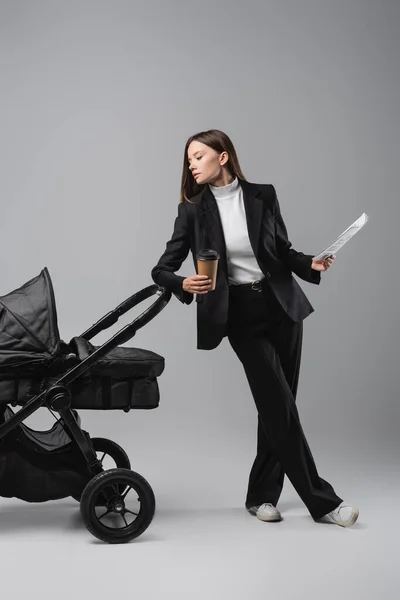 Young businesswoman standing with takeaway drink and newspaper near black pram on grey — Foto stock