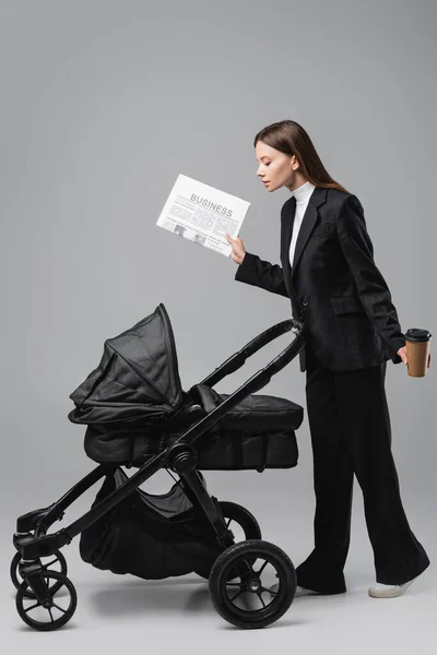 Vista lateral de la mujer en traje sosteniendo periódico y taza de papel cerca de cochecito negro en gris - foto de stock