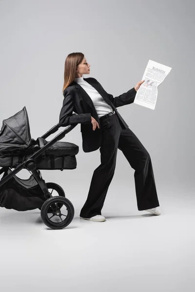 Full length view of woman reading newspaper while leaning on black pram on grey — Stock Photo