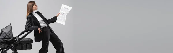 Young businesswoman reading newspaper near baby carriage isolated on grey, banner — Foto stock