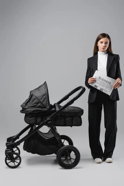 Woman in black suit standing with business newspaper near baby carriage on grey — Stock Photo