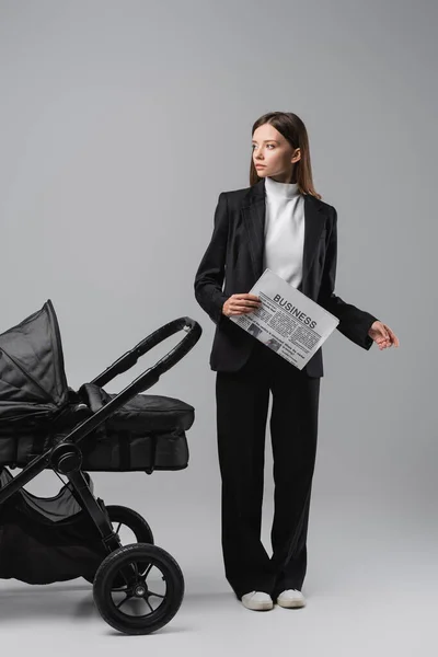 Full length view of young woman with business newspaper near baby carriage on grey — Foto stock