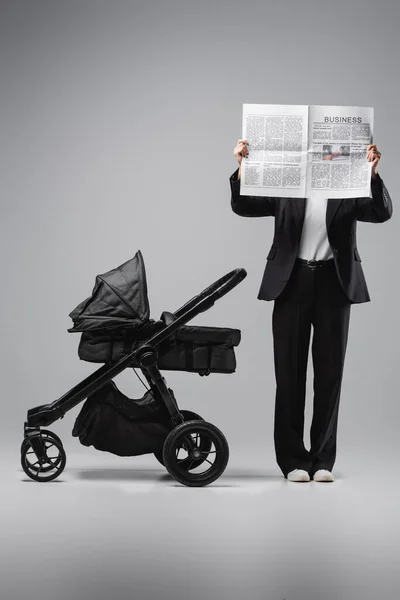 Mulher de terno preto obscurecendo rosto com jornal perto de carruagem do bebê em cinza — Fotografia de Stock