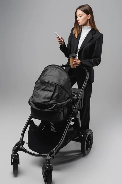 Mulher de negócios olhando para o smartphone enquanto está de pé com copo de papel perto de carrinho preto no cinza — Fotografia de Stock