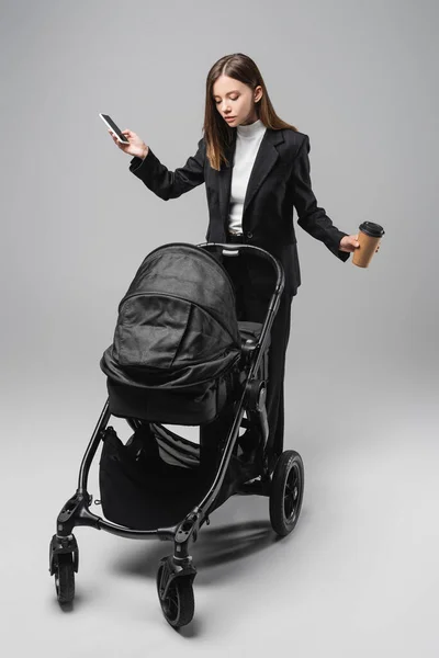 Businesswoman with takeaway drink and cellphone standing near black pram on grey — Stock Photo