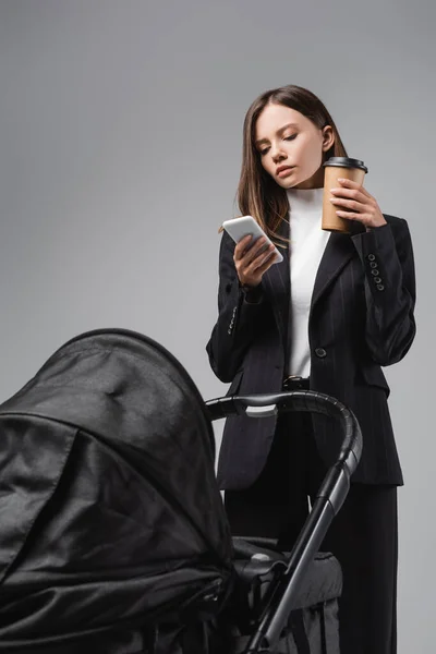 Young woman with coffee to go using mobile phone near black baby carriage isolated on grey — Stock Photo
