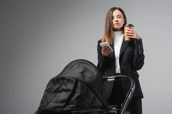 Young businesswoman with smartphone and coffee to go looking at camera near pram isolated on grey — Stock Photo