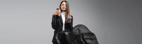 Frau mit Smartphone und Kaffee vor der Kamera in der Nähe von Kinderwagen isoliert auf grau, Banner — Stockfoto