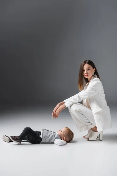 Mujer en traje blanco mirando a la cámara cerca de hijo acostado en gris - foto de stock