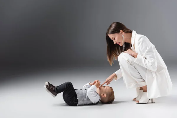 Side view of cheerful woman in white suit touching nose of son lying on grey — Stockfoto