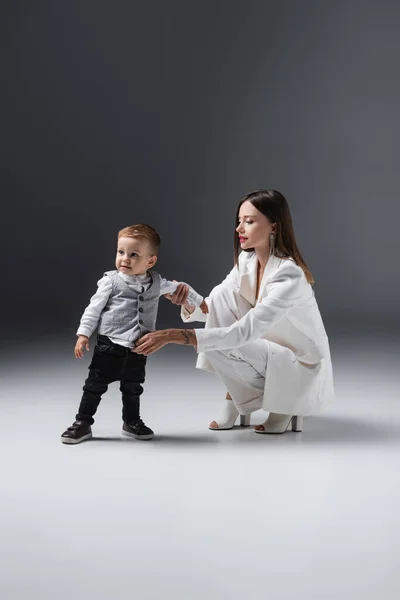 Mujer en traje blanco apoyando a su hijo pequeño de pie sobre gris - foto de stock