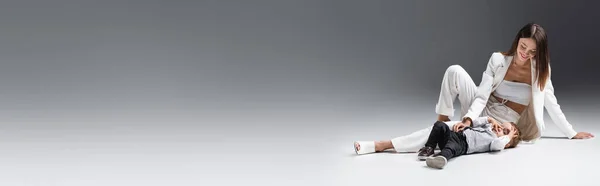 Pleased woman in white suit having fun with toddler boy lying on grey, banner - foto de stock