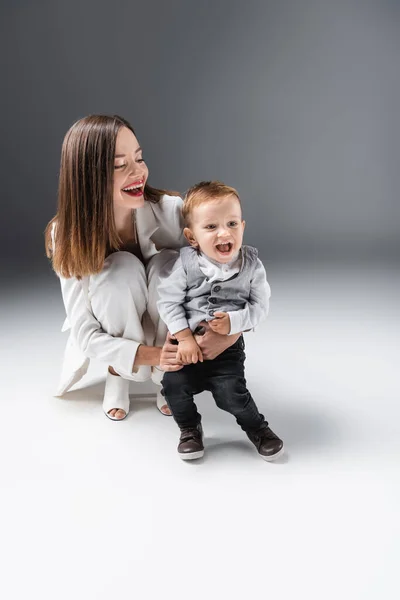 Femme heureuse embrassant fils étonné debout avec bouche ouverte sur gris — Photo de stock