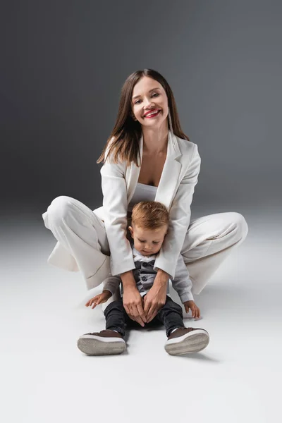 Веселая женщина смотрит в камеру и обнимает сына, сидящего на сером — стоковое фото