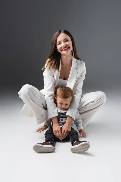 Cheerful woman looking at camera and embracing son sitting on grey — Fotografia de Stock