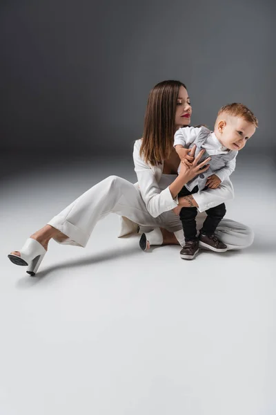 Ganzkörperansicht einer Frau im weißen Anzug, die ihren kleinen Sohn umarmt, während sie auf grau sitzt — Stockfoto