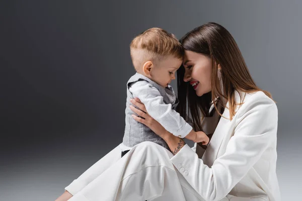 Fröhliche Frau im weißen Anzug mit kleinem Sohn auf grau — Stockfoto