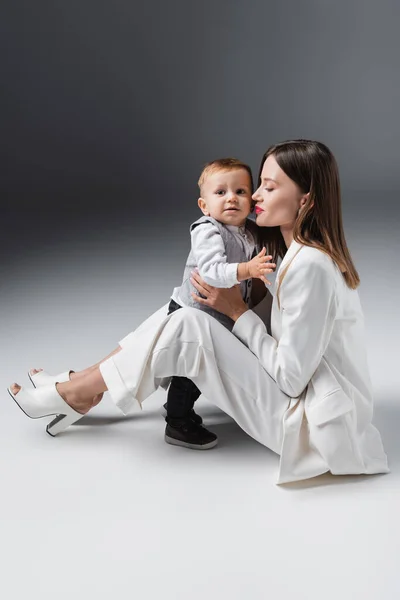 Повний вигляд жінки в білому костюмі, що обіймає сина, сидячи на сірому — стокове фото