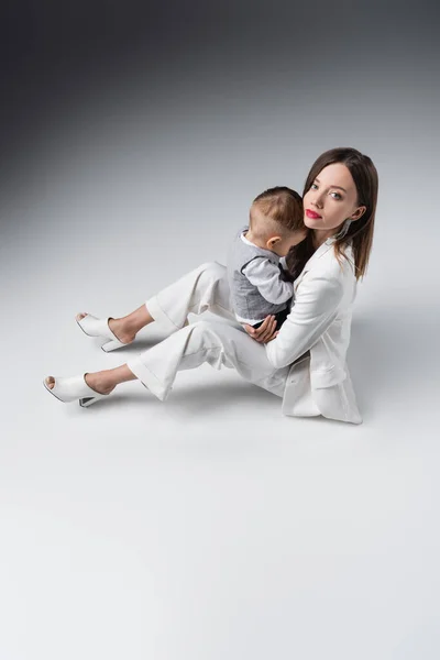 Vue grand angle de la femme à la mode regardant la caméra tout en étant assis avec enfant en bas âge fils sur gris — Photo de stock