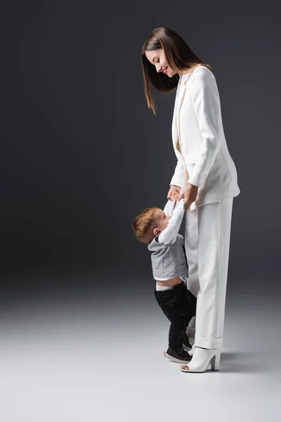 Full length view of stylish woman holding hands of son while having fun on grey — Fotografia de Stock