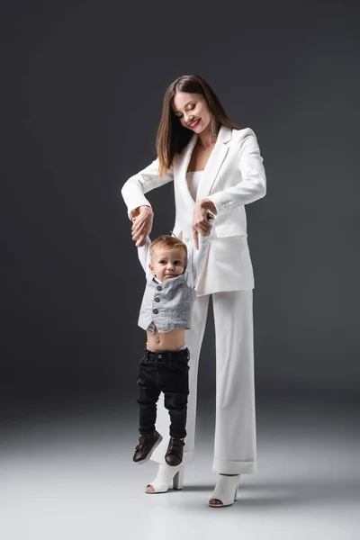 Sonriente mujer levantando pequeño hijo mientras sostiene sus manos en gris - foto de stock