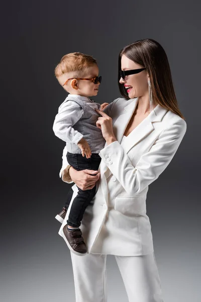 Щаслива мати і син в модному одязі і сонцезахисних окулярах дивиться один на одного на сірий — стокове фото