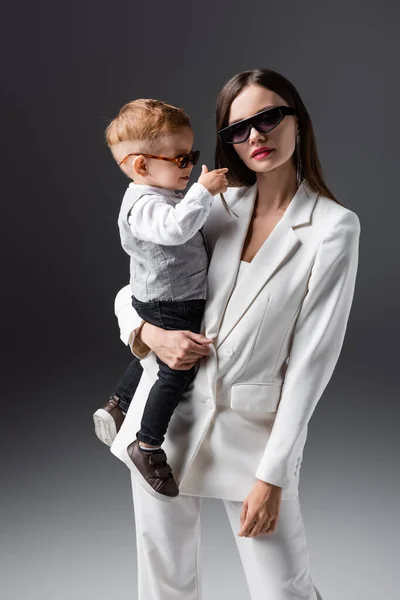 Trendy woman in sunglasses holding little son and looking at camera on grey — Stockfoto