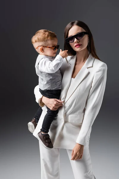 Modische Frau mit Sonnenbrille hält kleinen Sohn und blickt in Kamera auf grau — Stockfoto