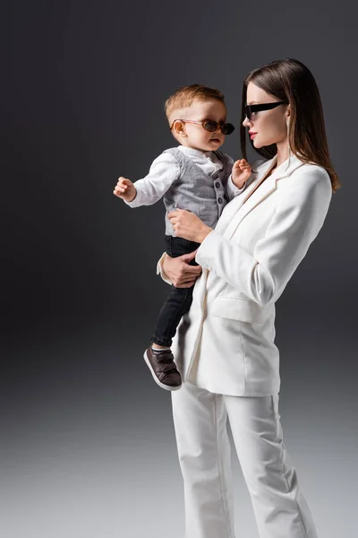 Trendy woman in suit and sunglasses holding baby boy on grey — Stockfoto
