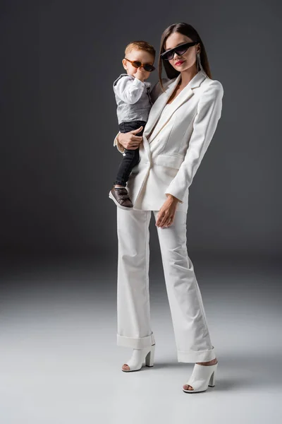Full length view of woman in trendy suit holding little son adjusting sunglasses on grey - foto de stock
