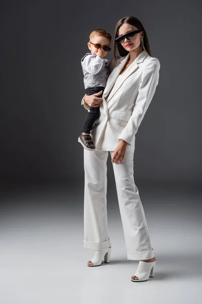 Vista completa de la mujer en traje de moda sosteniendo pequeño hijo ajustando gafas de sol en gris - foto de stock