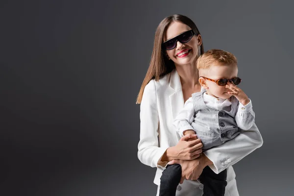 Happy woman in sunglasses and white blazer holding baby boy isolated on grey — Photo de stock