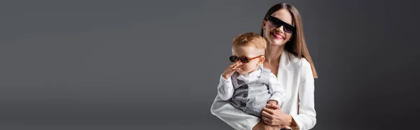 Pleased woman in white blazer holding son touching trendy sunglasses isolated on grey, banner — Stock Photo