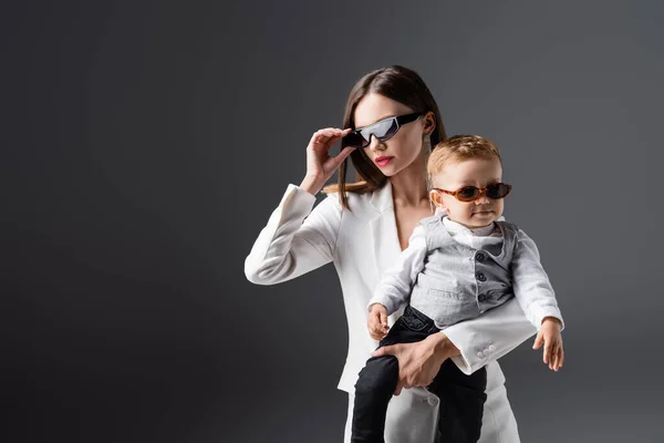 Joven mujer ajustando elegante gafas de sol mientras sostiene hijo aislado en gris - foto de stock