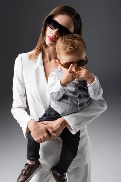 Stylish woman holding little son adjusting sunglasses on grey — стоковое фото