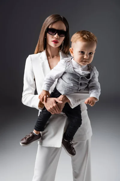 Женщина в солнечных очках держит сына в стильной одежде на сером — стоковое фото