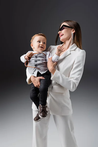 Laughing woman holding son wearing stylish clothes on grey - foto de stock
