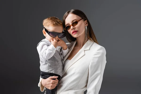 Mujer en gafas de sol y blazer blanco sosteniendo hijo pequeño aislado en gris - foto de stock