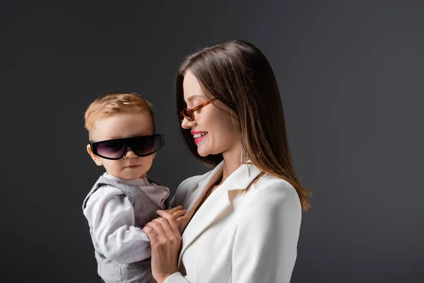 Niño en gafas de sol de moda mirando a la cámara cerca de mamá feliz aislado en gris - foto de stock