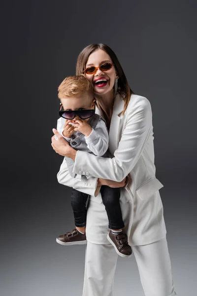 Laughing woman holding son adjusting stylish eyeglasses on grey — Stockfoto